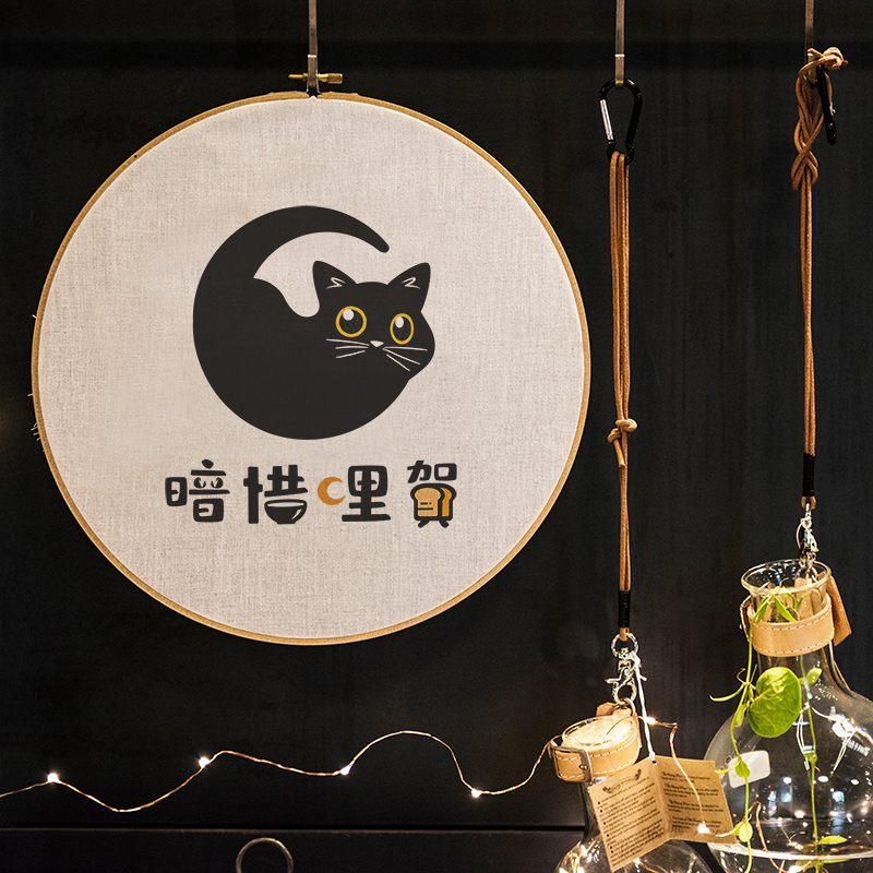圓形布面刺繡框中間為黑貓插畫 Logo，結合新月與大眼貓形象，下方為手寫風格字體『暗巷裡賀』，整體呈現溫馨療癒的宵夜品牌風格。